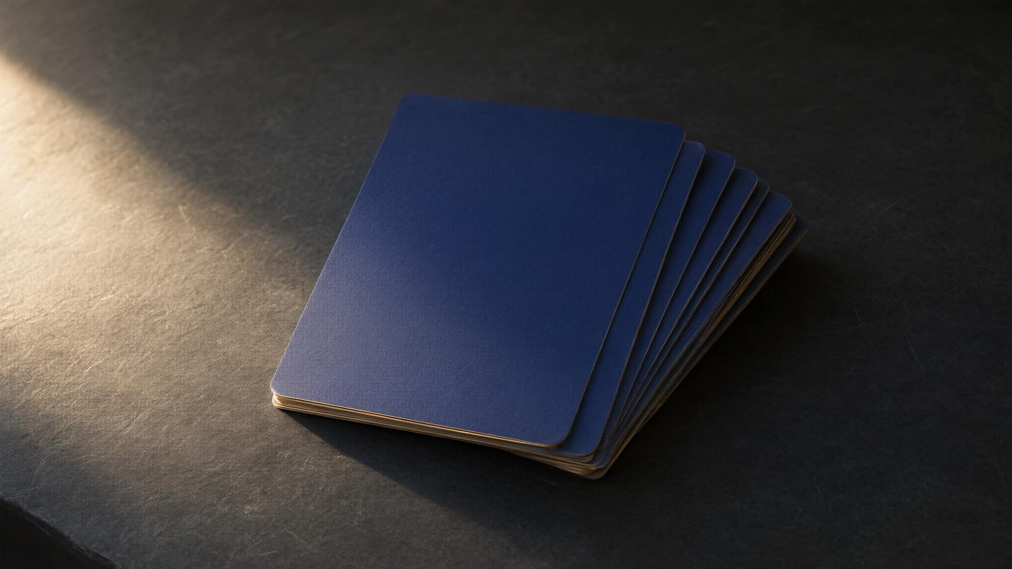 A small fanned stack of blank indigo cards with gold edges on dark stone, in a single raking light.