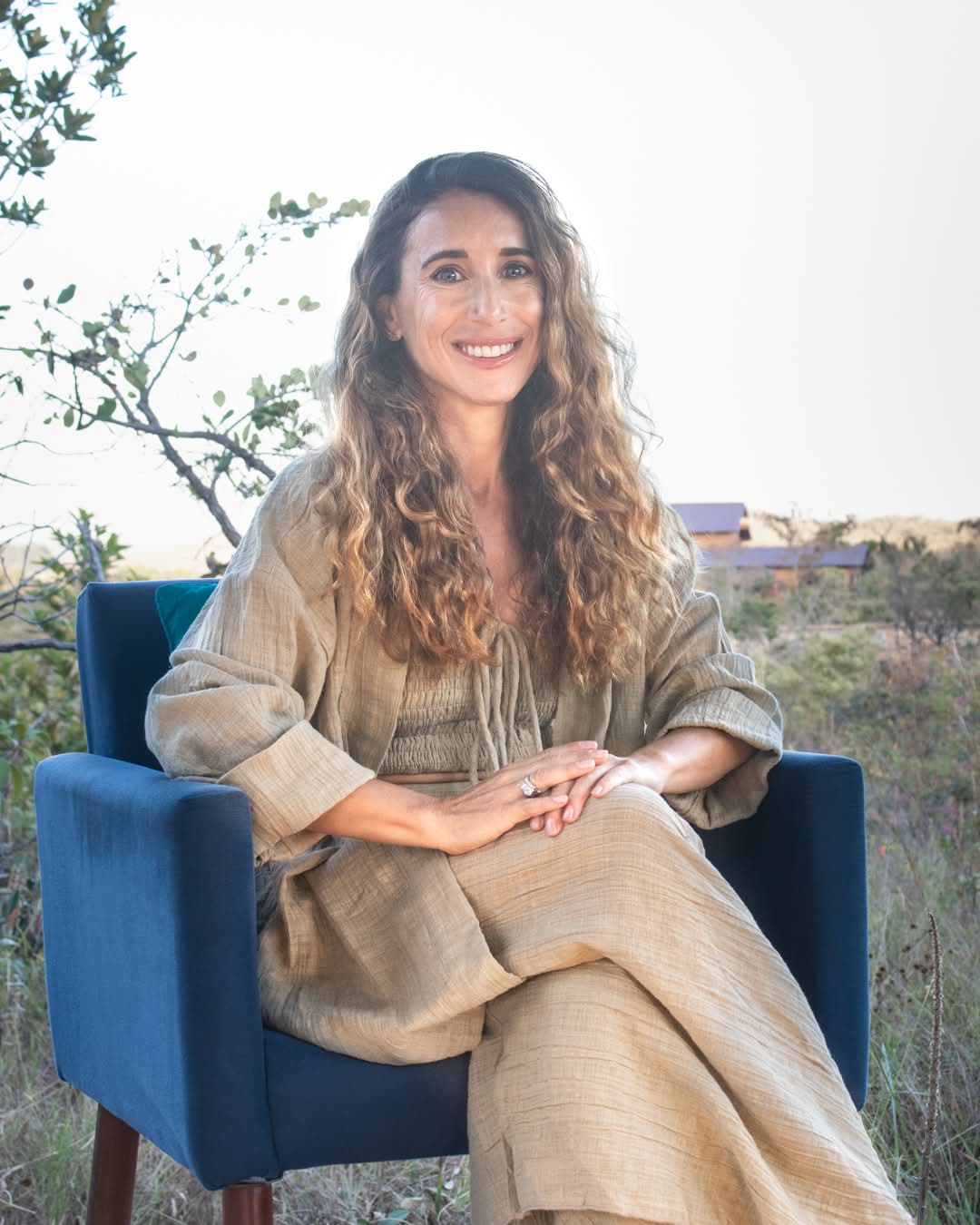 Jennifer, smiling, seated in a blue armchair outdoors in the late light, in a linen jacket, with long curling hair.
